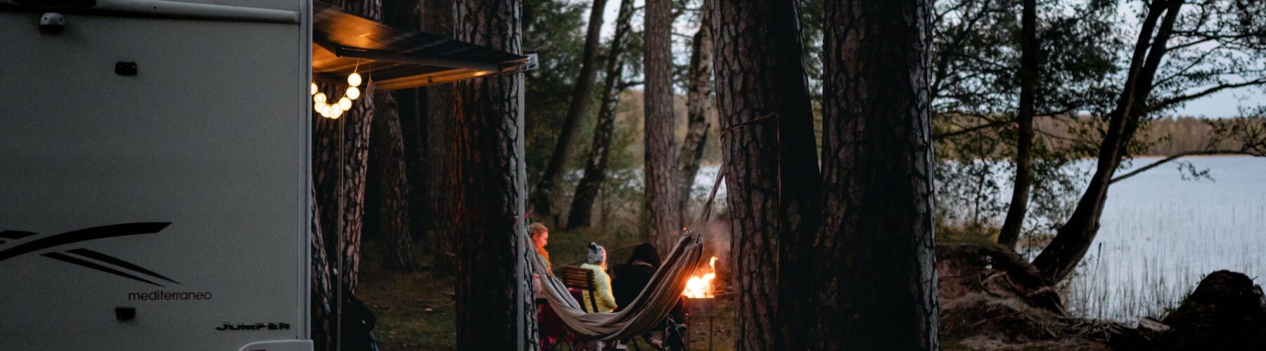 a couple sitting around a campfire outside of their camper which is situated on the shore of lake tahoe amongst the trees